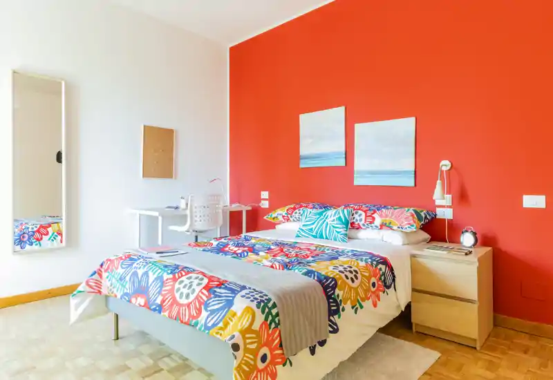 Camera da letto luminosa e colorata con letto matrimoniale e biancheria floreale vivace, comodino con lampada e sveglia, una scrivania e quadri su una parete accentuata di rosso.