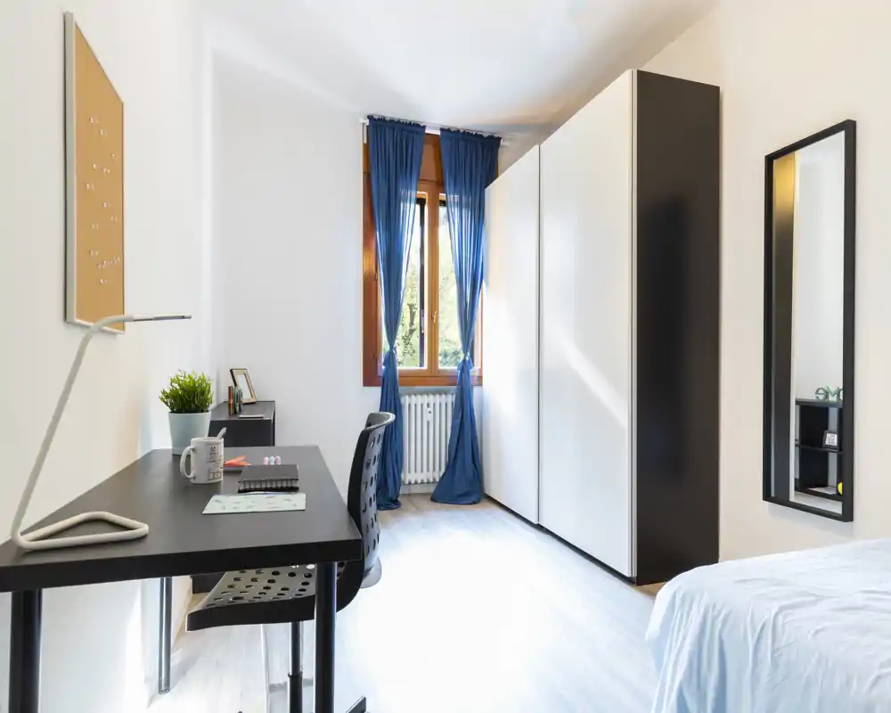 Bright, tidy bedroom with a visible single bed, a black desk and chair, large wardrobe and a window with blue curtains letting in natural light — ideal for a student or single occupant.