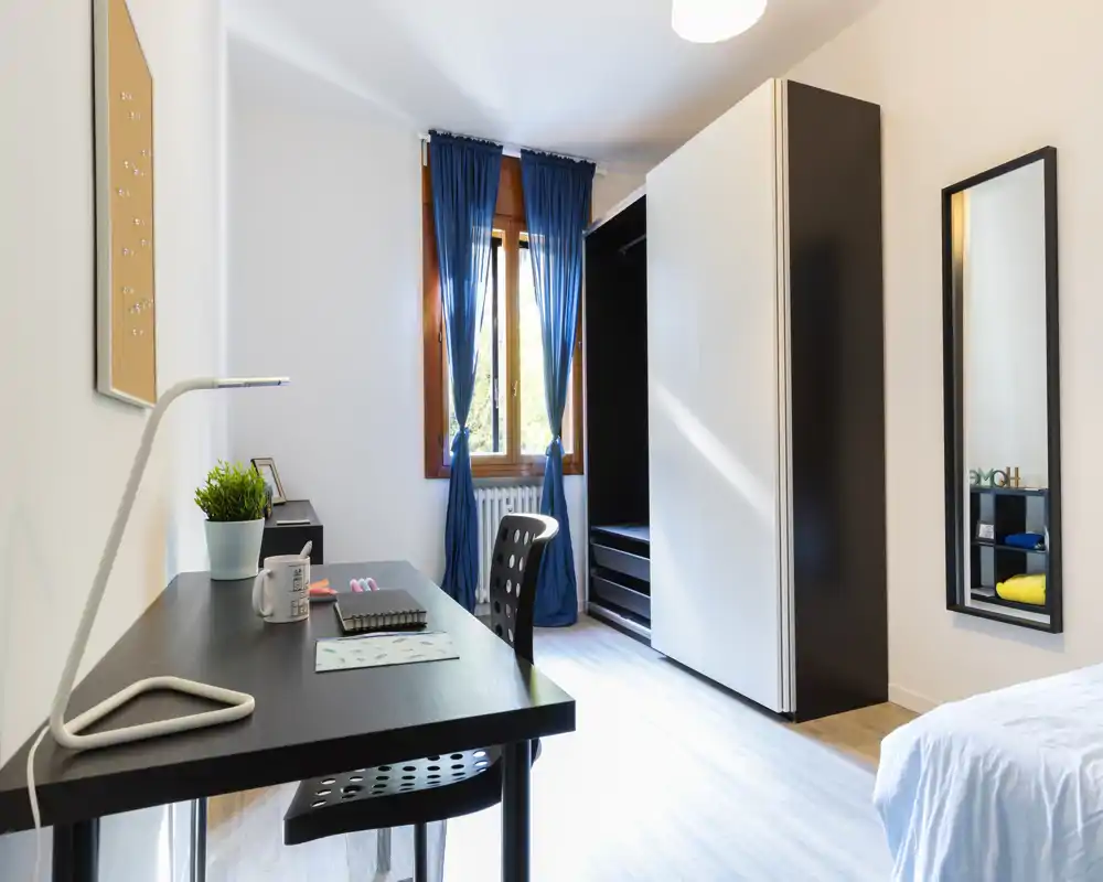 Bright, simply furnished bedroom with a study desk, wardrobe and a partially visible bed in the foreground. Deep blue curtains and wooden window frames provide contrast; the workspace includes a modern lamp and chair.