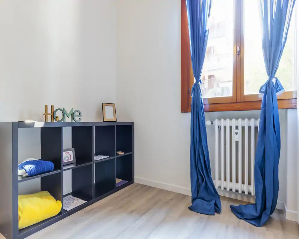 Bright small entrance or corner with a low storage cubby, wooden floor and a window with blue curtains letting in natural light.