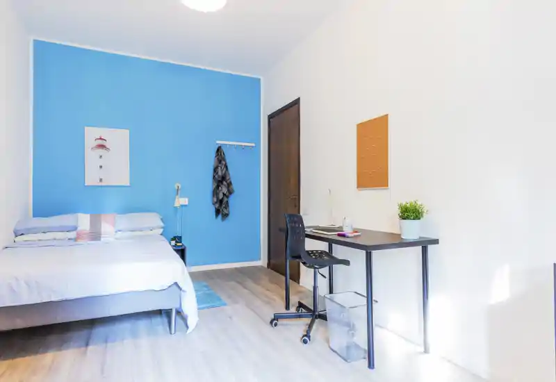 Bright, modern bedroom with a double bed against a vivid blue accent wall, bedside table and a workspace with desk and chair. Natural light fills the room, creating an airy and welcoming atmosphere ideal for short-term rentals.