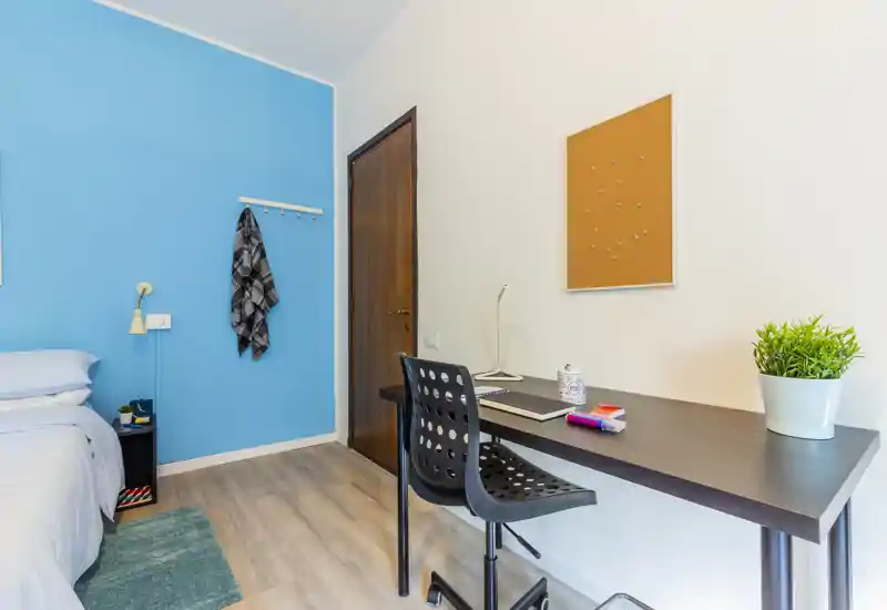 Bright, modern bedroom with a blue accent wall, a neatly made bed on the left and a functional desk with chair on the right — ideal for a student or remote worker.