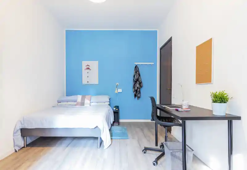 Bright, modern bedroom with a full bed against an eye-catching blue accent wall, bedside table and a compact workspace with a desk and chair — ideal for a short-term rental or student flat.