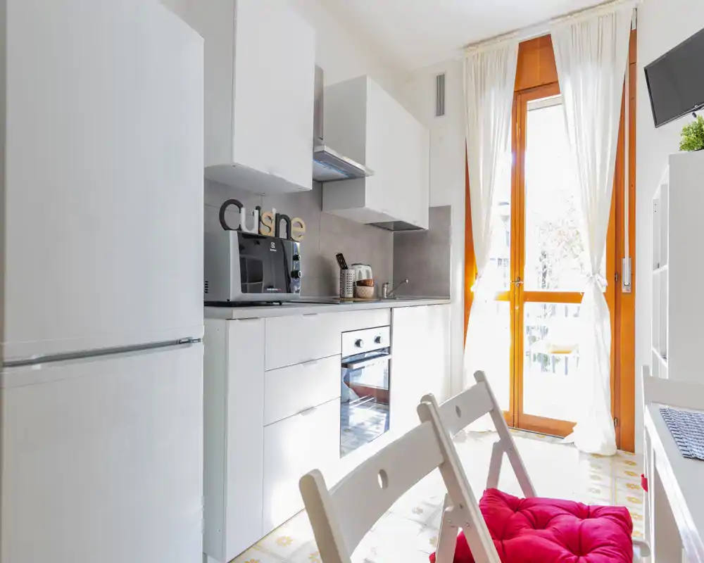 Bright, modern kitchenette with white cabinets, built-in oven, fridge and a sunny glass door leading outside. Simple, well-styled space ideal for short-term rentals.