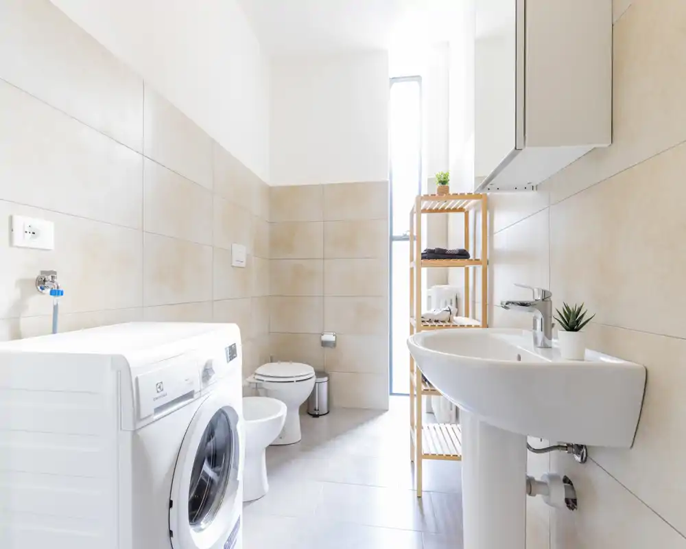 Bright modern bathroom with a pedestal sink, toilet, bidet and a front-loading washing machine; neutral tiles and a slim tall window bring in natural light.