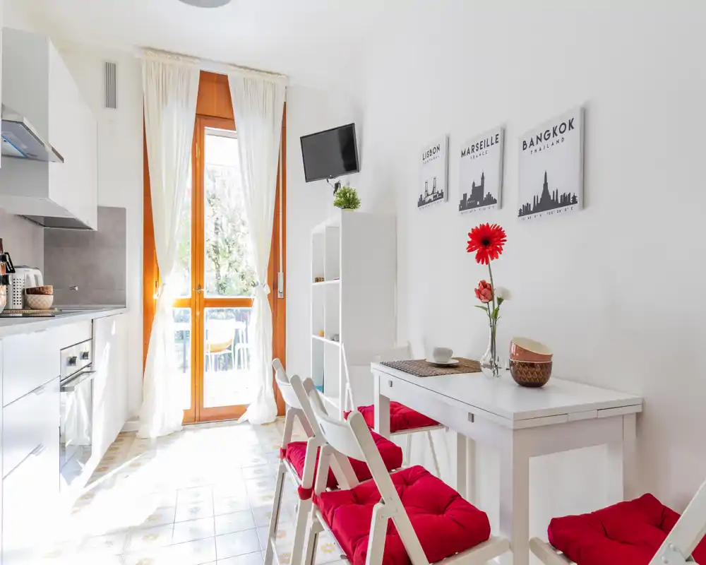 Bright, compact kitchen with white cabinetry, a wood-framed glass door leading to a balcony, and a small dining area with red-cushioned chairs — modern and well-lit.