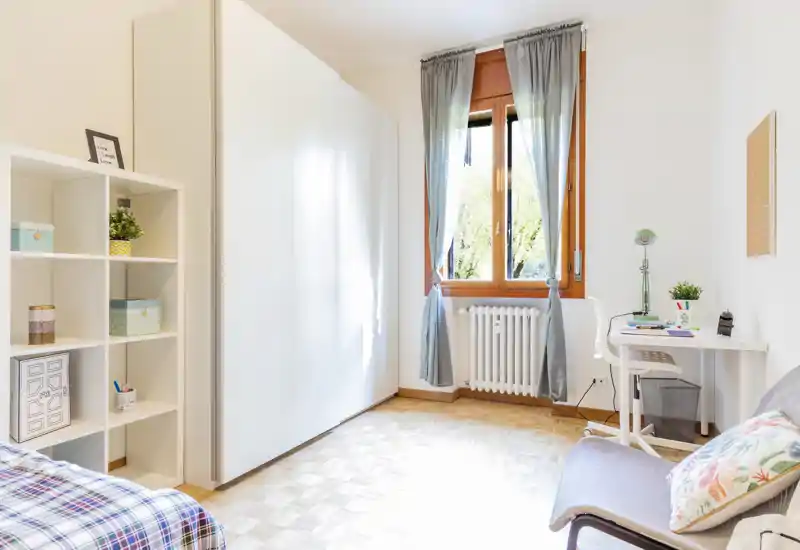 Bright, airy bedroom with a partial bed view, white wardrobe, shelving and a small work desk by the window — ideal for a single occupant.