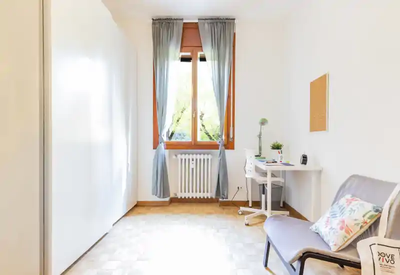 Bright, minimal living area with a comfortable chair, decorative cushion and a small desk by the window; parquet flooring and soft curtains create a calm, airy atmosphere.
