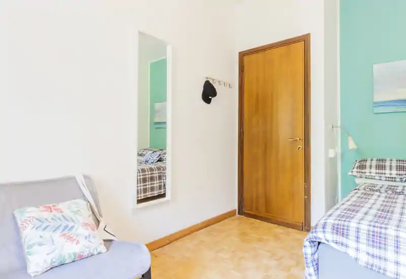 Sunny, neatly staged bedroom corner with a single bed dressed in checked linens, a bedside lamp, decorative painting and a mirror reflecting part of the bed. Light wooden floor and teal accent wall create a fresh, inviting atmosphere.