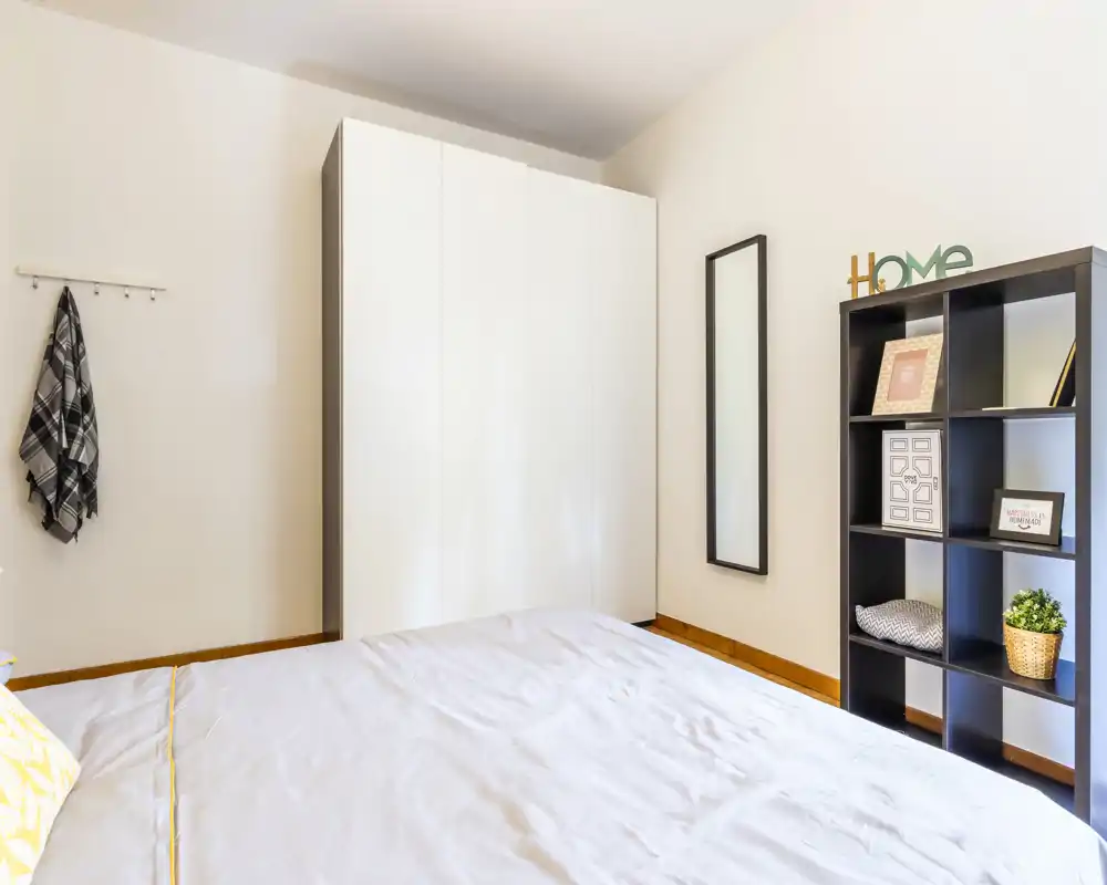 Camera da letto luminosa e minimal con un ampio letto visibile, armadio bianco e libreria con elementi decorativi — ambiente pulito e arioso ideale per soggiorni riposanti.