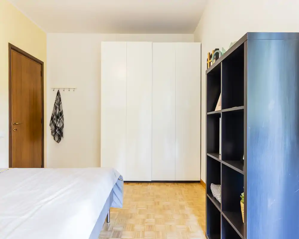 Camera da letto luminosa e arredata in modo essenziale con letto matrimoniale visibile, grande armadio bianco e scaffalatura aperta. Toni neutri e pavimento in parquet creano un’atmosfera pulita e tranquilla.