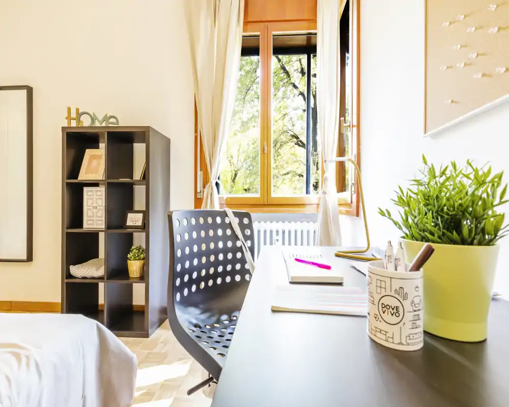 Angolo camera soleggiato e ben allestito con piccolo spazio lavoro: scrivania vicino a una grande finestra, sedia moderna traforata, scaffale e letto parzialmente visibile — luce naturale intensa e arredo fresco.