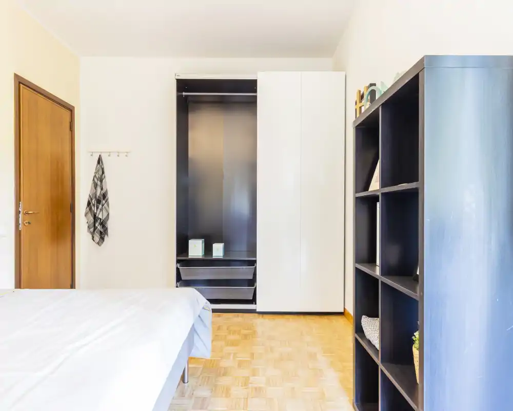 Camera da letto luminosa e minimale con letto ben rifatto, armadio aperto e libreria; parquet chiaro e pareti neutre creano un'atmosfera calma e ariosa.