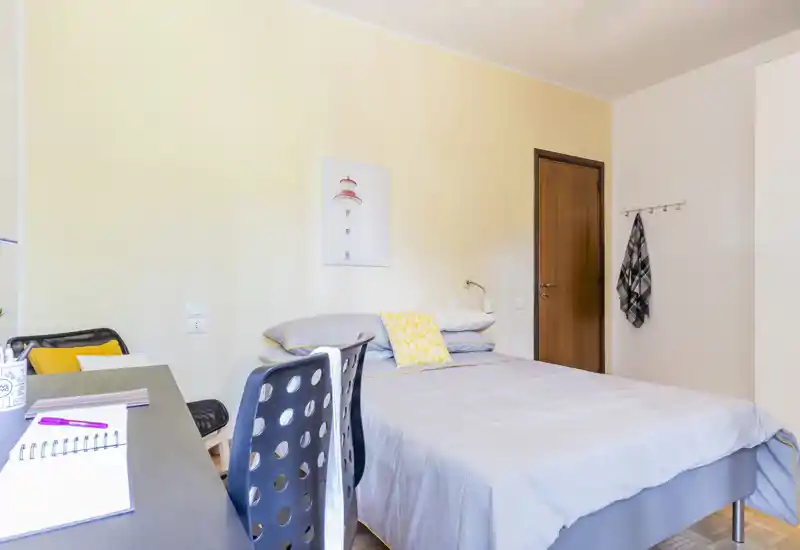 Camera da letto luminosa e ordinata con letto matrimoniale, biancheria grigia e cuscini gialli; una scrivania con sedia accanto al letto crea un pratico spazio di lavoro.