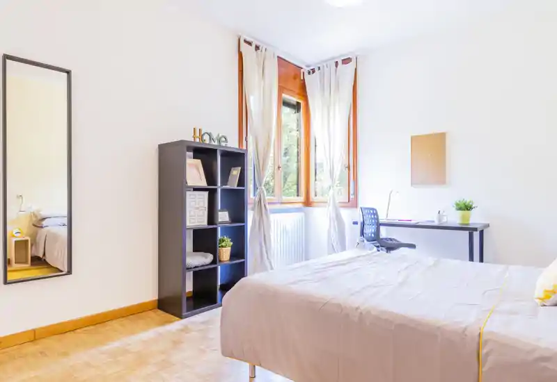 Camera da letto luminosa e minimal con letto matrimoniale ben rifatto, una scrivania vicino alla finestra e una libreria a cubi. La grande finestra con tende leggere illumina l'ambiente creando un'atmosfera fresca e ariosa, ideale per lavoro e riposo.