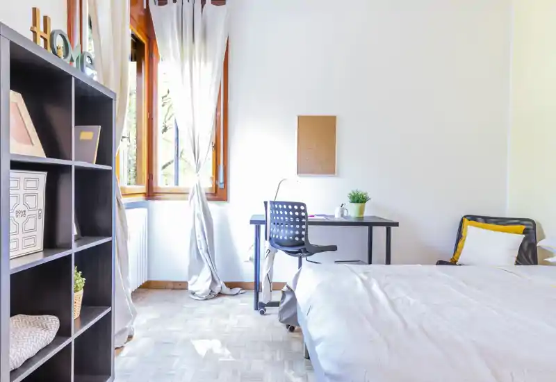 Camera da letto luminosa e ben arredata con letto comodo, scrivania vicino alla finestra, scaffalatura e luce naturale calda—ideale per riposo o lavoro da remoto.