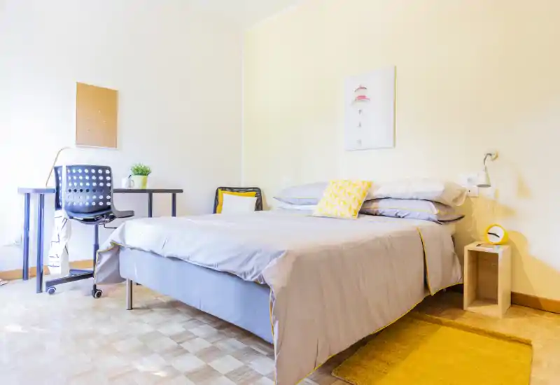 Camera da letto luminosa e curata con letto matrimoniale rifatto, biancheria grigia e tocchi gialli; una scrivania vicino alla finestra aggiunge un'area lavoro funzionale.