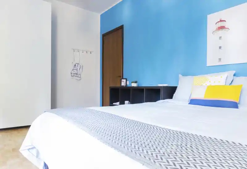 Bright, modern bedroom with a large bed in the foreground, colorful pillows and a blue accent wall creating a fresh, welcoming atmosphere.