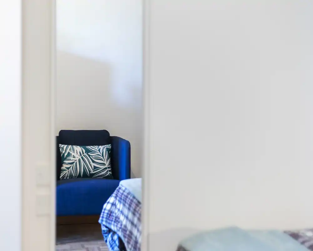 Cozy bedroom detail with a patterned cushion on a blue chair visible through a doorway and a portion of the bed in the foreground. Bright, sharply focused and suitable for highlighting corner seating and bedding textures.