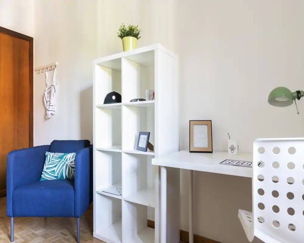 Bright, modern living corner with a blue armchair, white shelving unit and a tidy workspace — ideal for relaxing or light home office use.