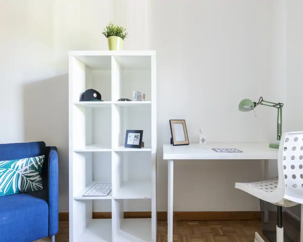 A bright, modern living area with a compact white shelving unit, a small desk and a blue sofa accented with a patterned cushion—clean, minimal and well lit.