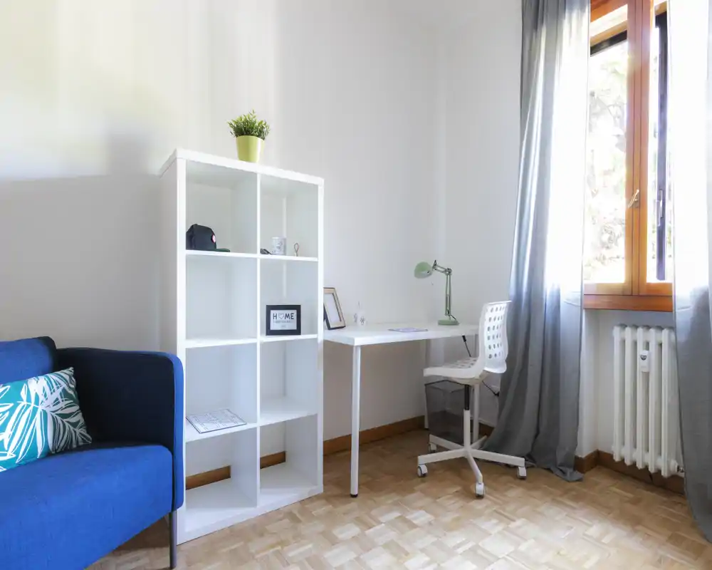 Bright, modern living area with a blue sofa, open shelving and a compact desk by a window — ideal for a cozy studio setup with natural light.