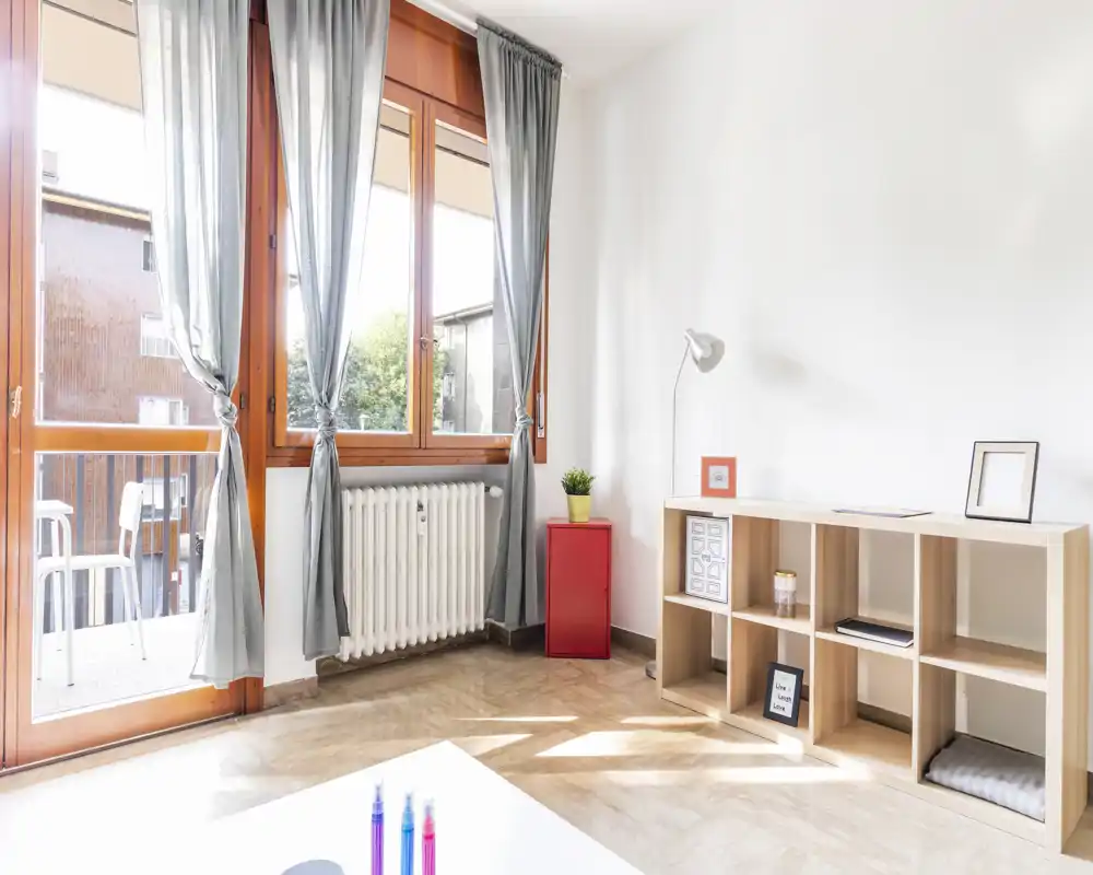 Zona giorno luminosa e minimal con ampia finestra e accesso al balcone. Mobile in legno chiaro, una piccola pianta e decorazioni semplici creano un'atmosfera pulita e ariosa.