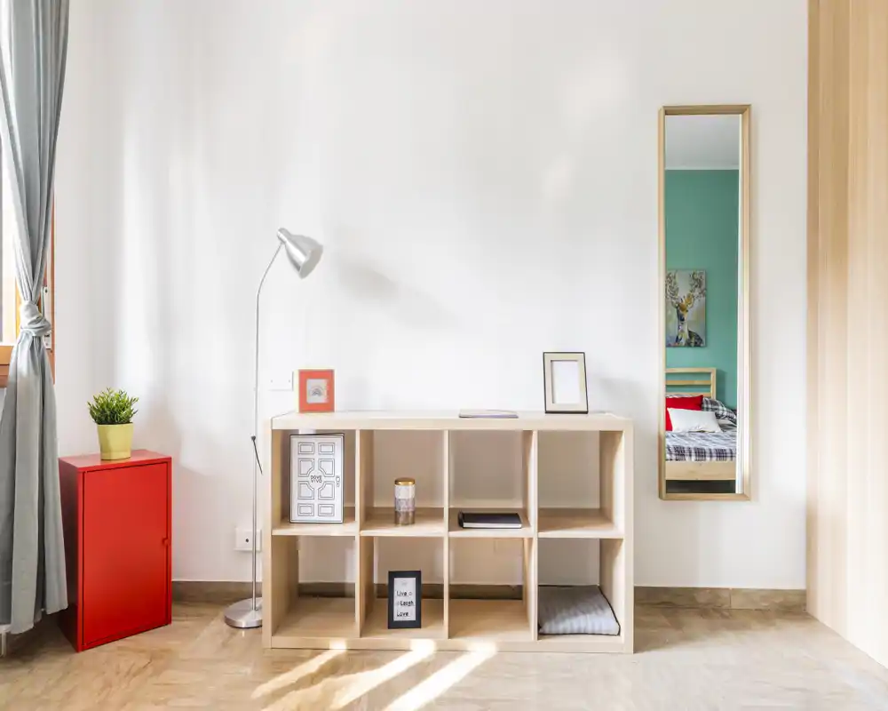Angolo di camera luminoso e minimale con un mobile a cubi, lampada da terra, piccolo armadietto rosso e uno specchio a figura intera che riflette un letto. Lo spazio è pulito e decorato con piante e cornici, ideale per un annuncio moderno.