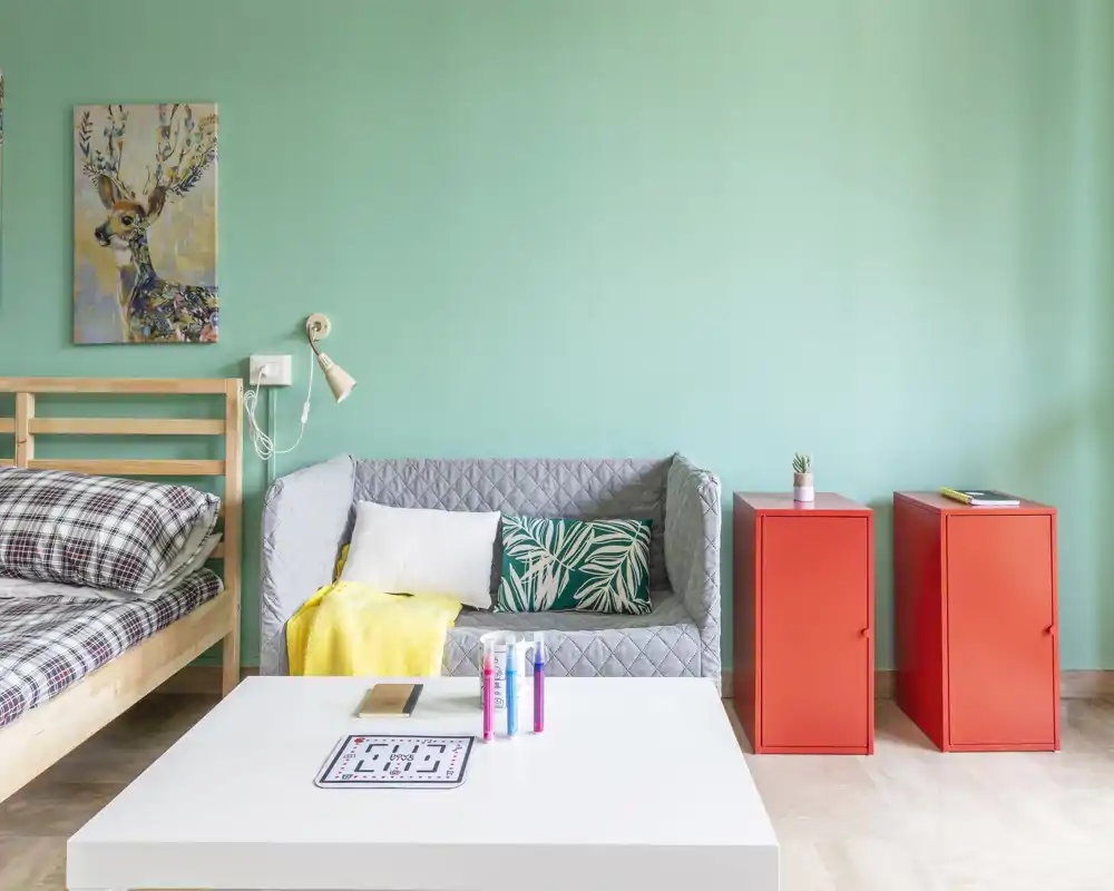 Angolo camera luminoso e accogliente con letto in legno, biancheria a quadri e un piccolo divano. La parete verde menta, il quadro colorato e i mobili rossi creano un aspetto moderno e fresco.