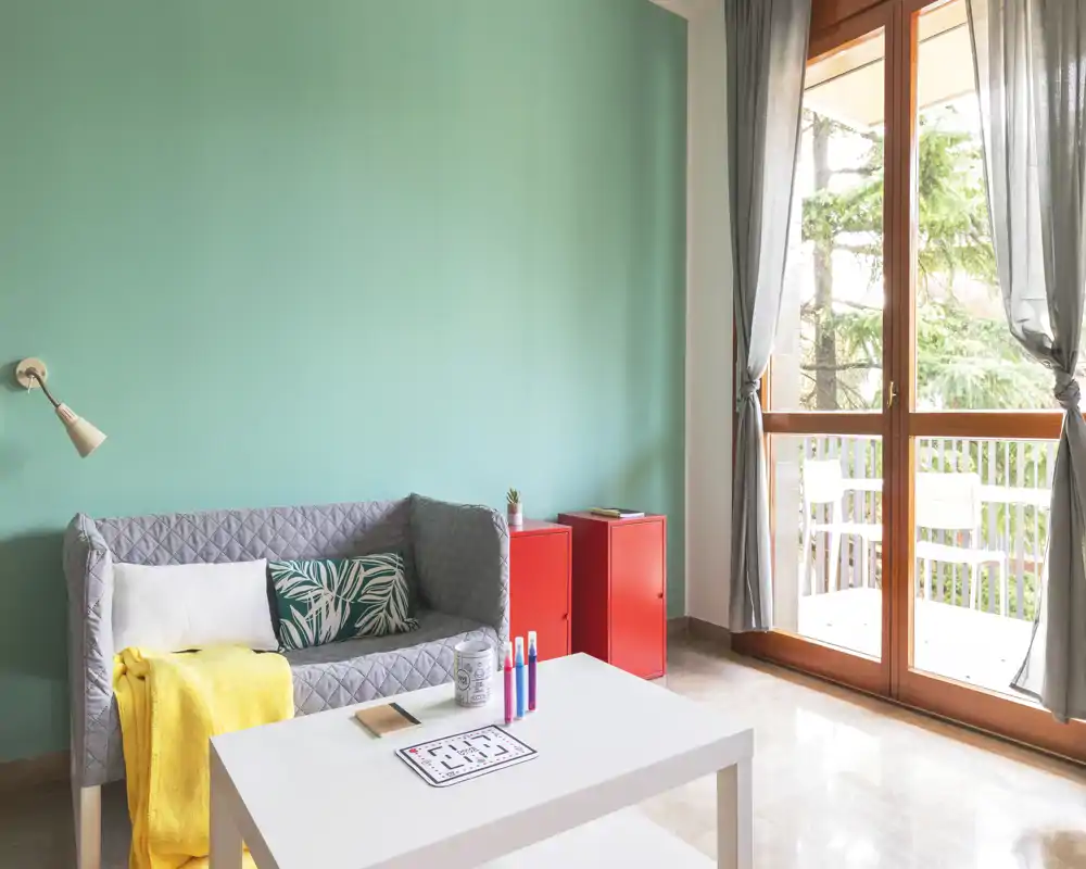 Zona giorno luminosa e accogliente con un divanetto compatto, cuscini decorativi e tavolino, con una grande porta sul balcone che illumina l’ambiente.