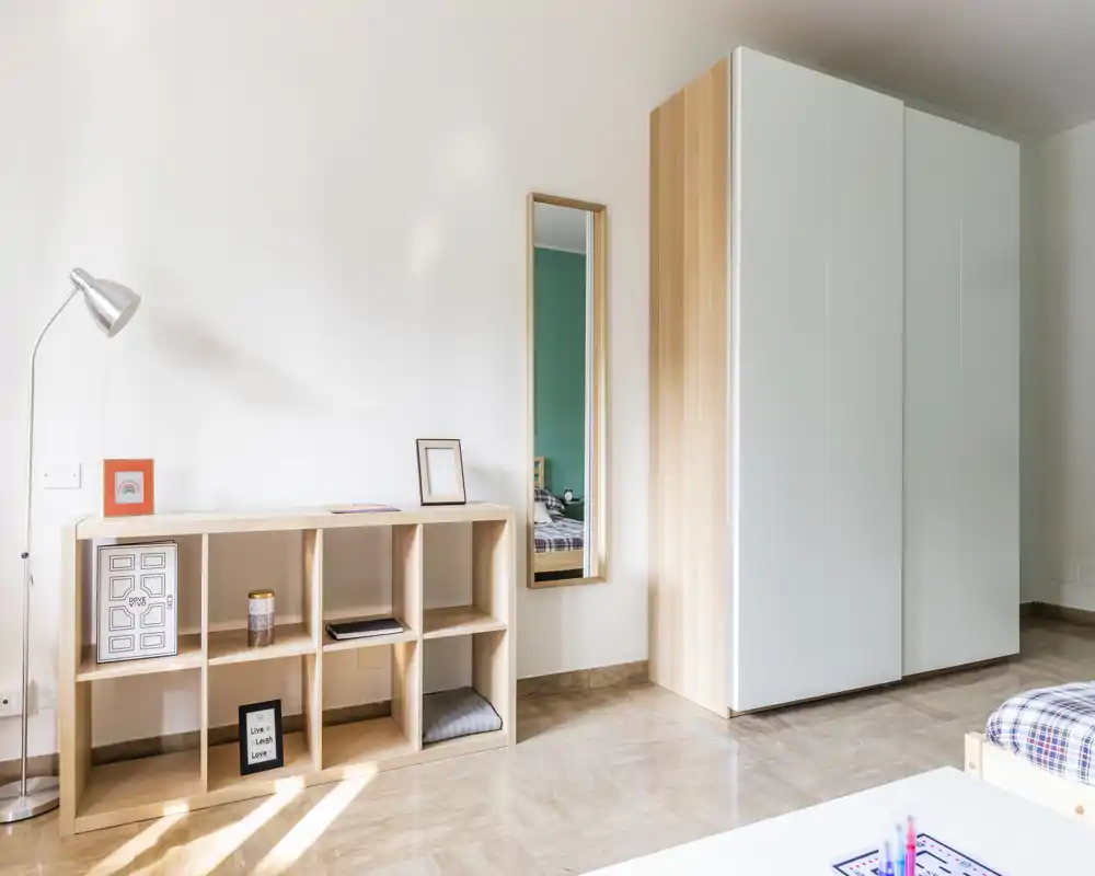 Angolo di camera luminoso e minimal con armadio, scaffalatura e vista parziale del letto; linee pulite, luce naturale e arredo semplice.