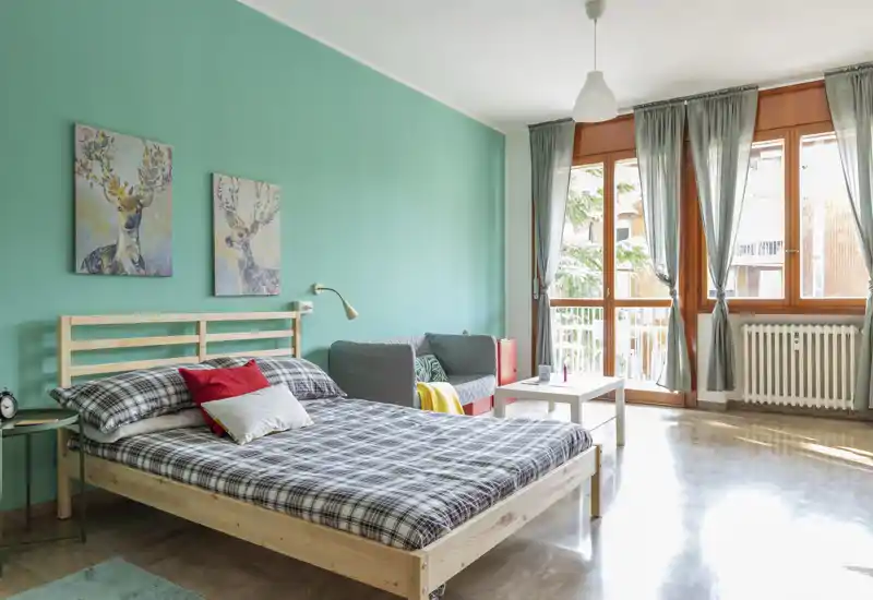 Bright, stylish bedroom with a wooden bed frame, patterned bedding and colorful accent pillows. Large windows flood the room with natural light and a small seating area with a grey sofa and coffee table completes the layout.