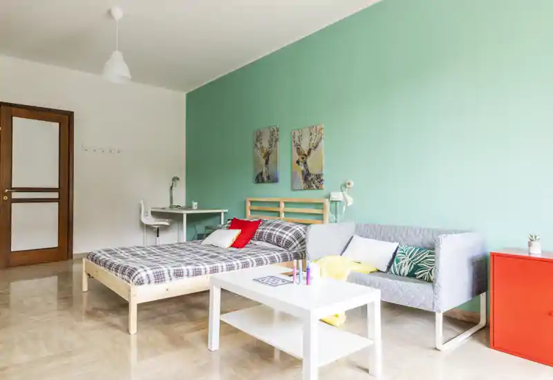 Bright, modern bedroom with a wooden bed, patterned bedding and a small lounge area with a grey sofa and coffee table against a mint-green accent wall.