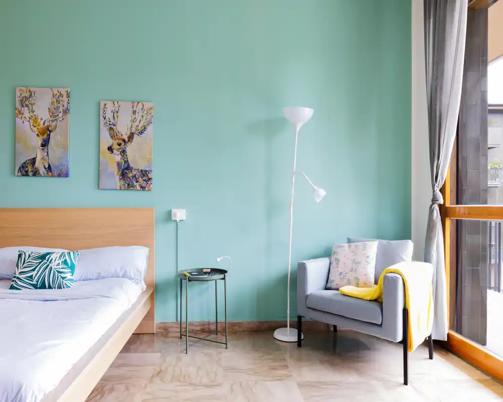 Bright, modern bedroom with a wooden platform bed, patterned cushions, colorful deer artwork and a cozy armchair next to a large window letting in natural light.
