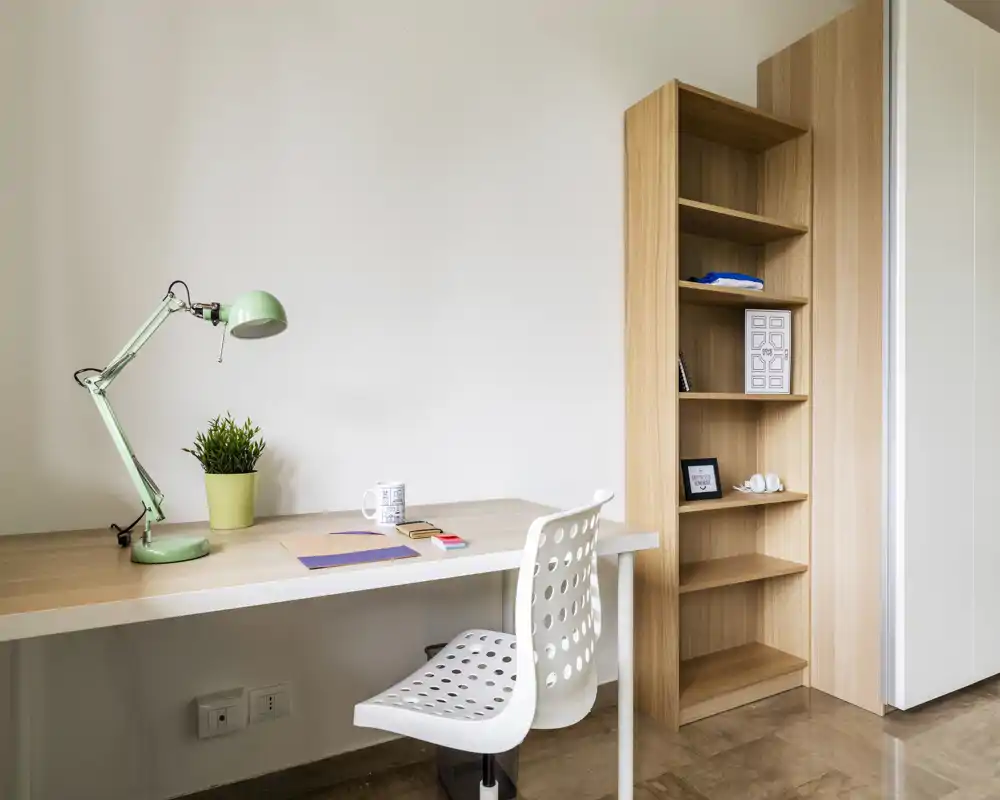 Bright, minimalist workspace corner with a wooden desk, perforated white chair, adjustable green desk lamp and open shelving — ideal for focused study or remote work.