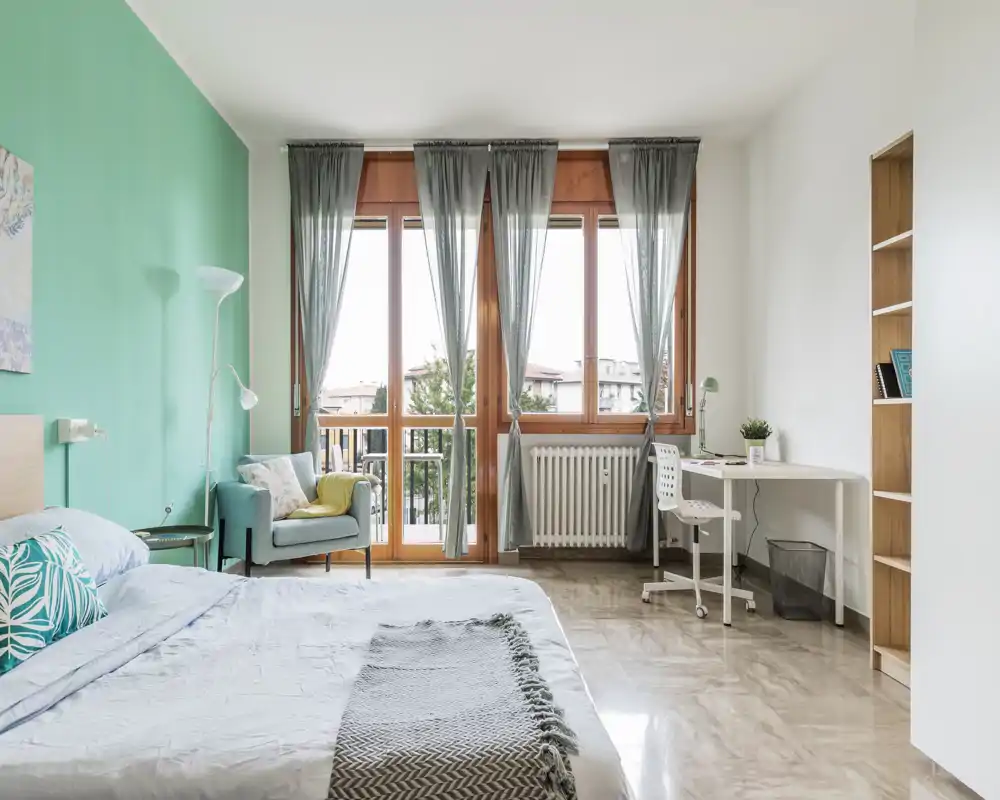 Bright, stylish bedroom with a large bed in the foreground, teal accent wall, comfy armchair and a study corner by the window letting in abundant natural light.