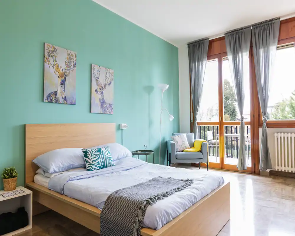 Bright, stylish bedroom with a wooden bedframe, teal accent wall, matching artwork and a large window/door letting in abundant natural light. The room includes a cozy armchair and soft textiles that create a welcoming atmosphere.