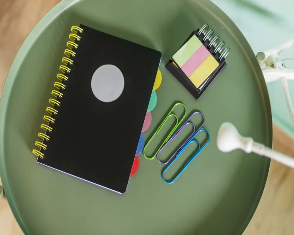 Close-up of a small round surface with stationery: a spiral notebook, sticky notes and oversized colorful paperclips next to a desk lamp. The photo focuses on desk accessories rather than showing a room.