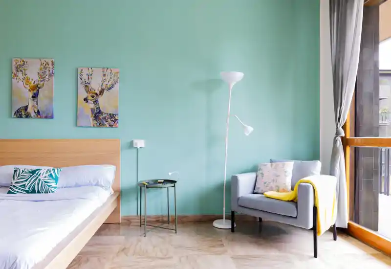 Bright, modern bedroom with a wooden platform bed, patterned cushions, colorful deer artwork and a cozy armchair next to a large window letting in natural light.