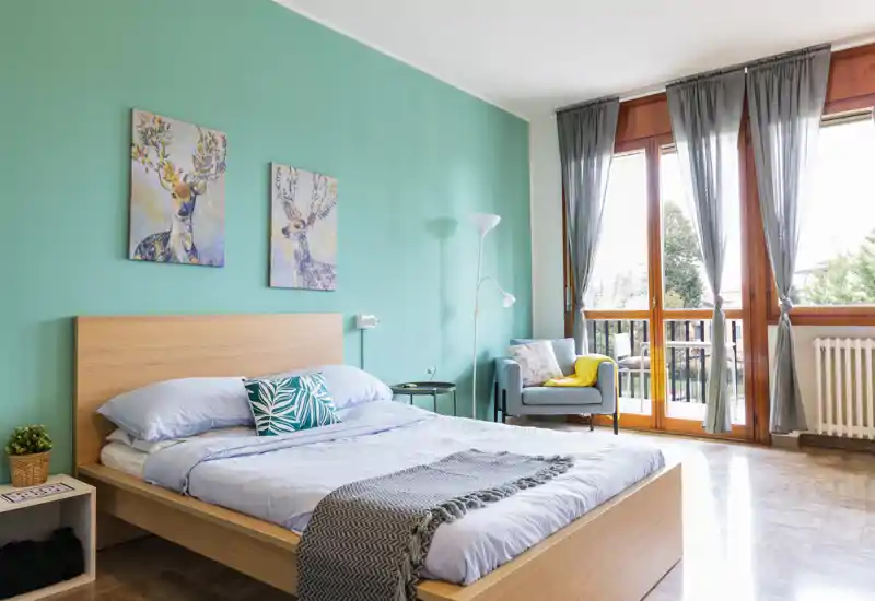 Bright, stylish bedroom with a wooden bedframe, teal accent wall, matching artwork and a large window/door letting in abundant natural light. The room includes a cozy armchair and soft textiles that create a welcoming atmosphere.