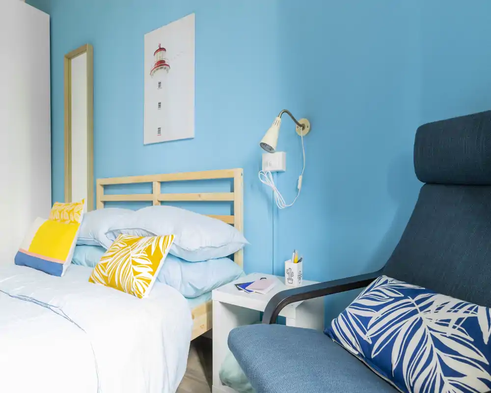 Bright, cozy bedroom with a light wood bedframe, layered pale-blue bedding and colorful accent pillows; a comfortable armchair and bedside table complete the composition.