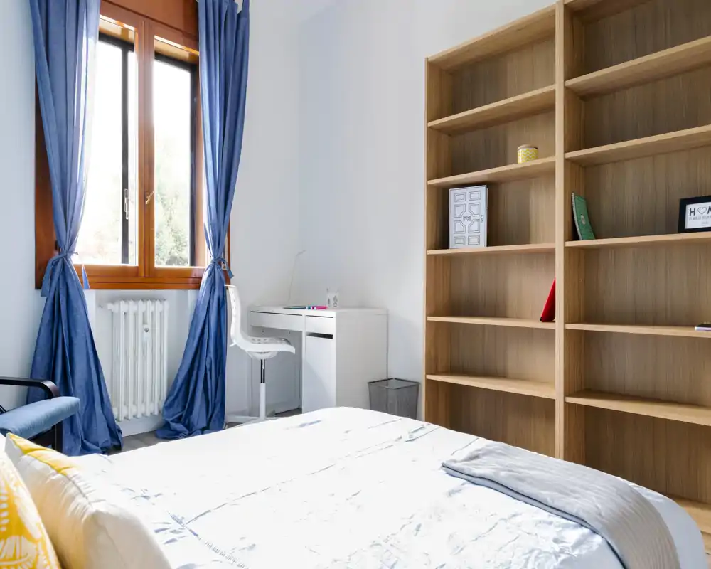 Bright, airy bedroom with a neatly made bed, large window with blue curtains, a small desk and tall wooden shelving — ideal for a comfortable stay.