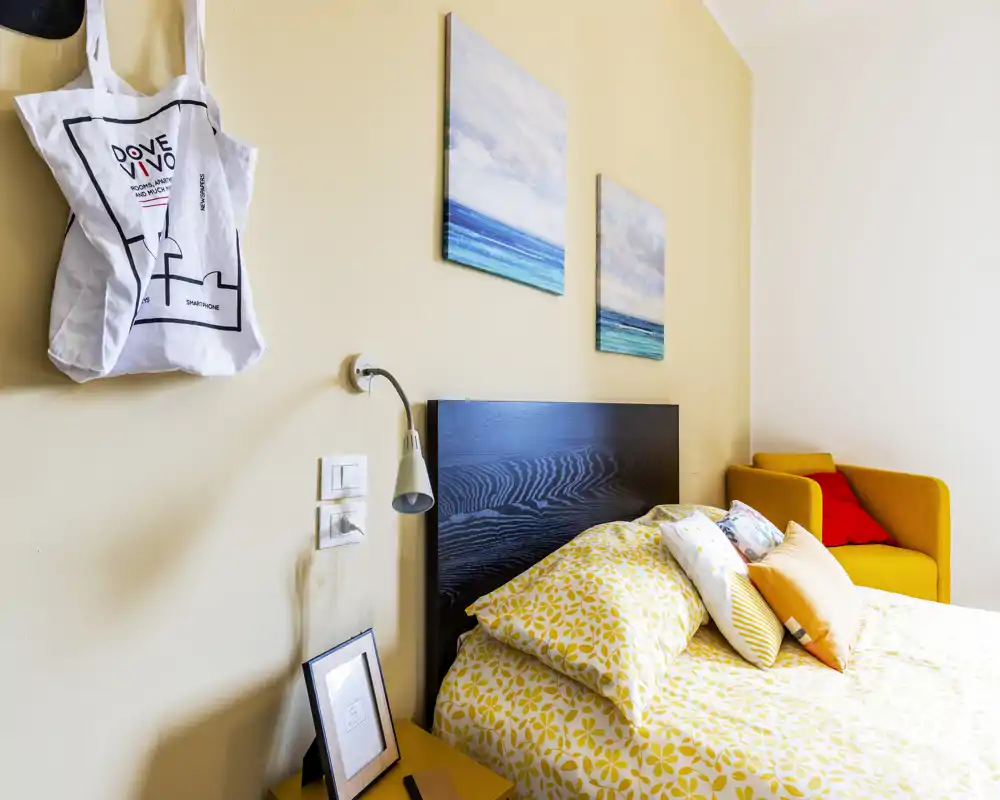Bright, neatly styled bedroom featuring a patterned yellow duvet, decorative pillows and a dark wooden headboard. A mustard armchair and coastal prints add color and a cozy atmosphere.