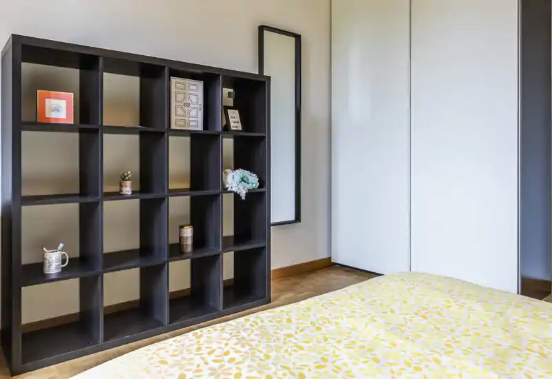 Bright, modern bedroom corner featuring a patterned yellow bedspread, an open cubed shelving unit and a tall mirror — clean, minimalist styling with warm wooden flooring.