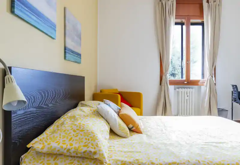 Bright, cozy bedroom with a large bed dressed in yellow patterned linens, decorative pillows and a warm accent armchair by the window letting in natural light.