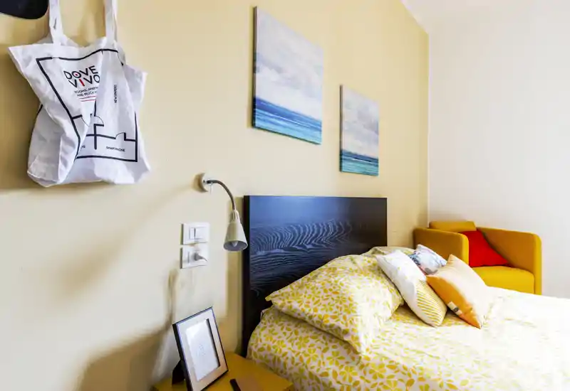 Bright, neatly styled bedroom featuring a patterned yellow duvet, decorative pillows and a dark wooden headboard. A mustard armchair and coastal prints add color and a cozy atmosphere.