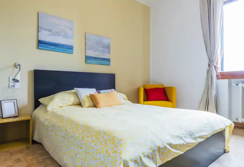 Bright, cozy bedroom with a large double bed, decorative cushions, wall art and a comfortable armchair by the window — ideal for a relaxing stay.
