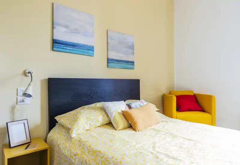 Bright, cozy bedroom with a patterned yellow duvet, decorative pillows and a dark wood headboard; a mustard armchair with a red cushion sits in the corner, and two seascape paintings hang above the bed.