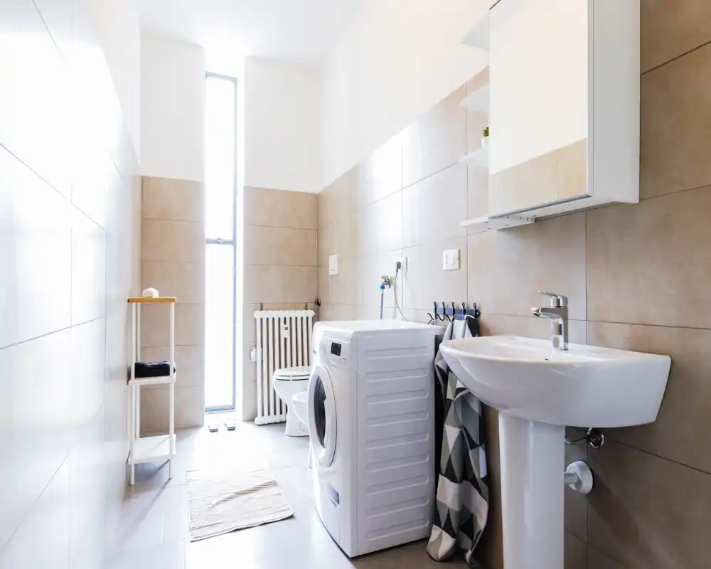 Bright, modern bathroom with pedestal sink, washing machine and toilet. Neutral tiles and tall narrow window provide strong natural light and a clean, functional layout ideal for listings.