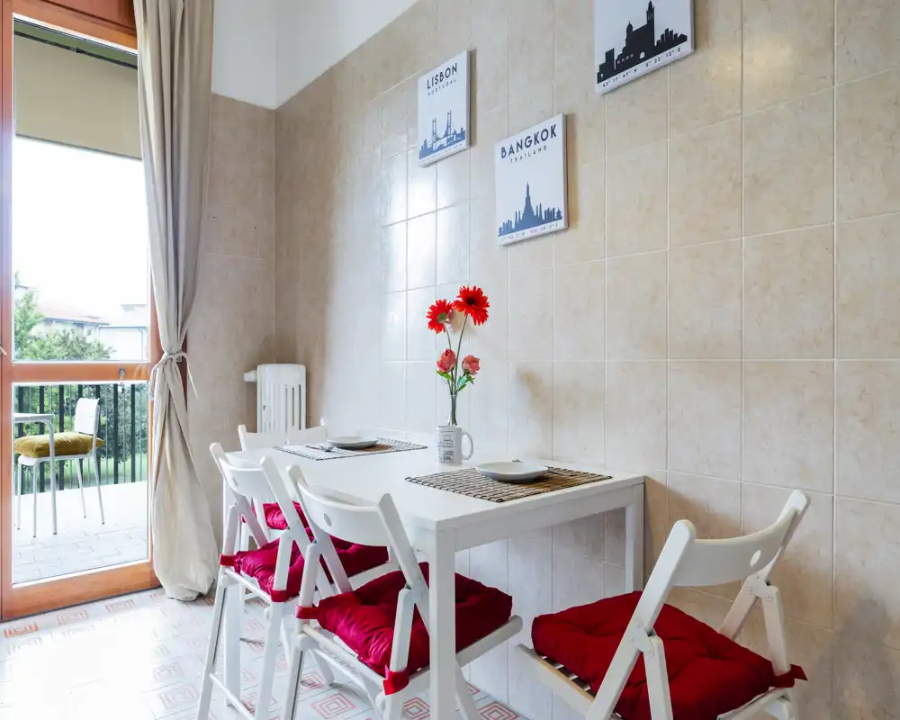 Bright, welcoming dining corner with a white table, red-cushioned chairs and a balcony view. Simple decorative touches — placemats and fresh flowers — create a cozy atmosphere.